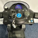 Approved Used Pride Go Go Elite Traveller in grey, with 12 ah batteries and mesh basket, front controls sitting on a navy blue carpet, behind is a white brick wall.