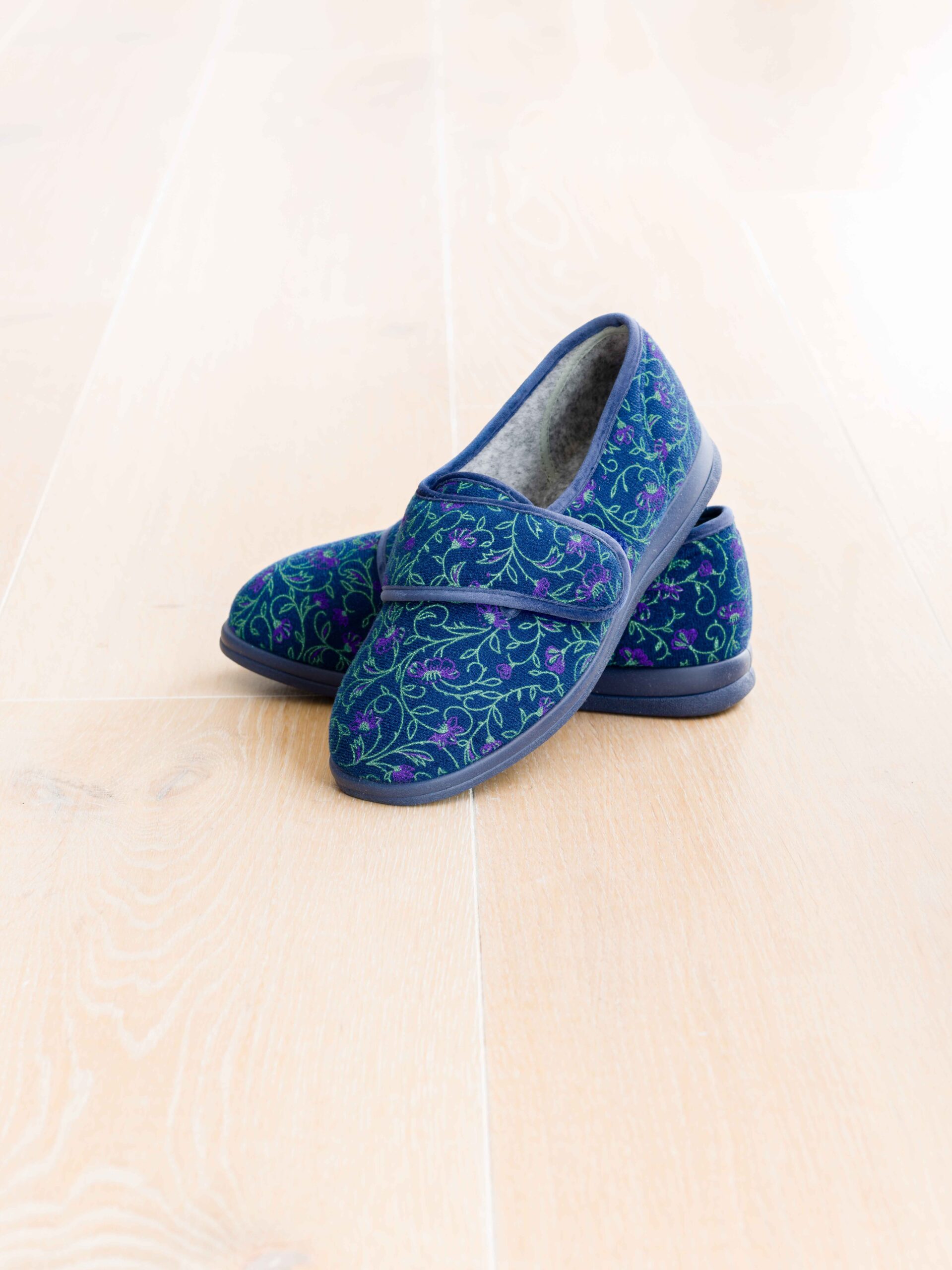 Blue floral-patterned Cosyfeet Holly slippers with purple accents are placed on a light wood floor, conveying a cozy and inviting atmosphere.