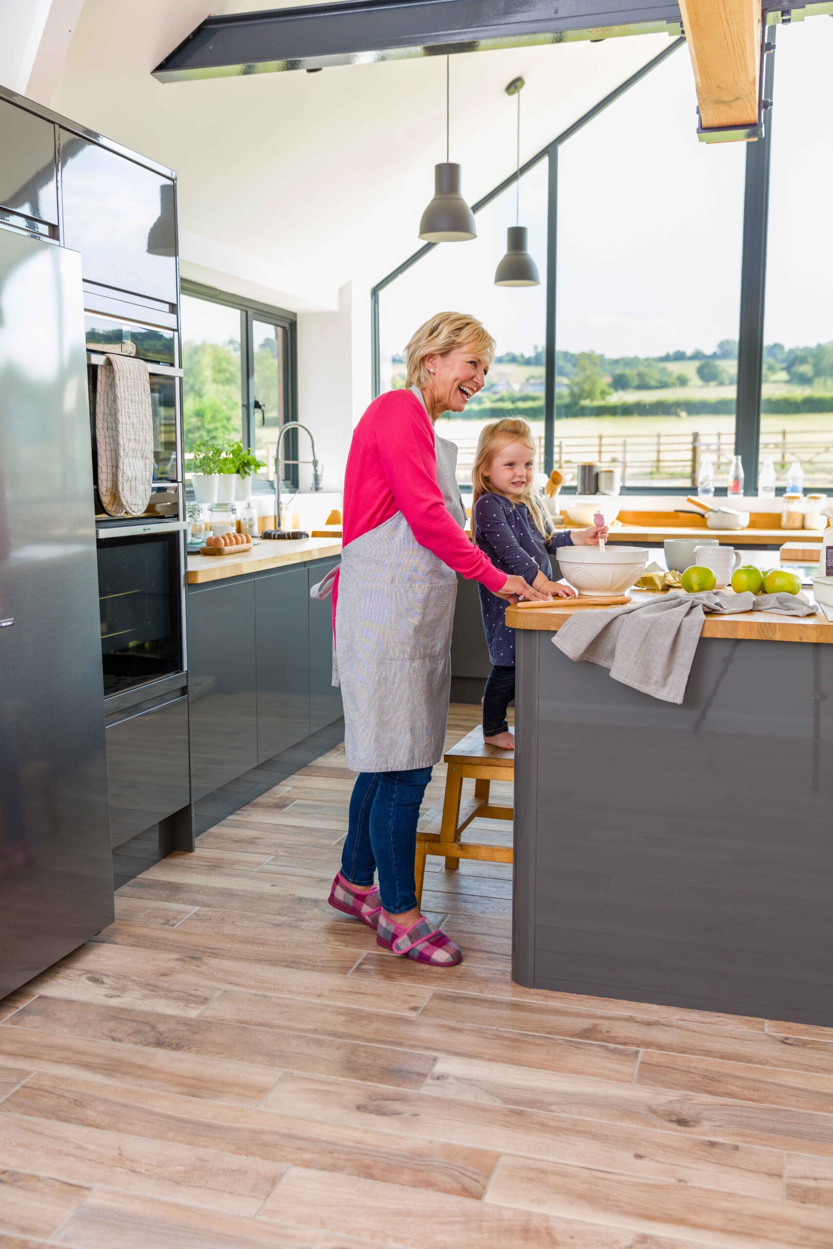 A woman wearing Cosyfeet Holly Slippers and a young girl in a bright, modern kitchen baking together. They are smiling by a counter with mixing bowls, green apples, and wooden stools.