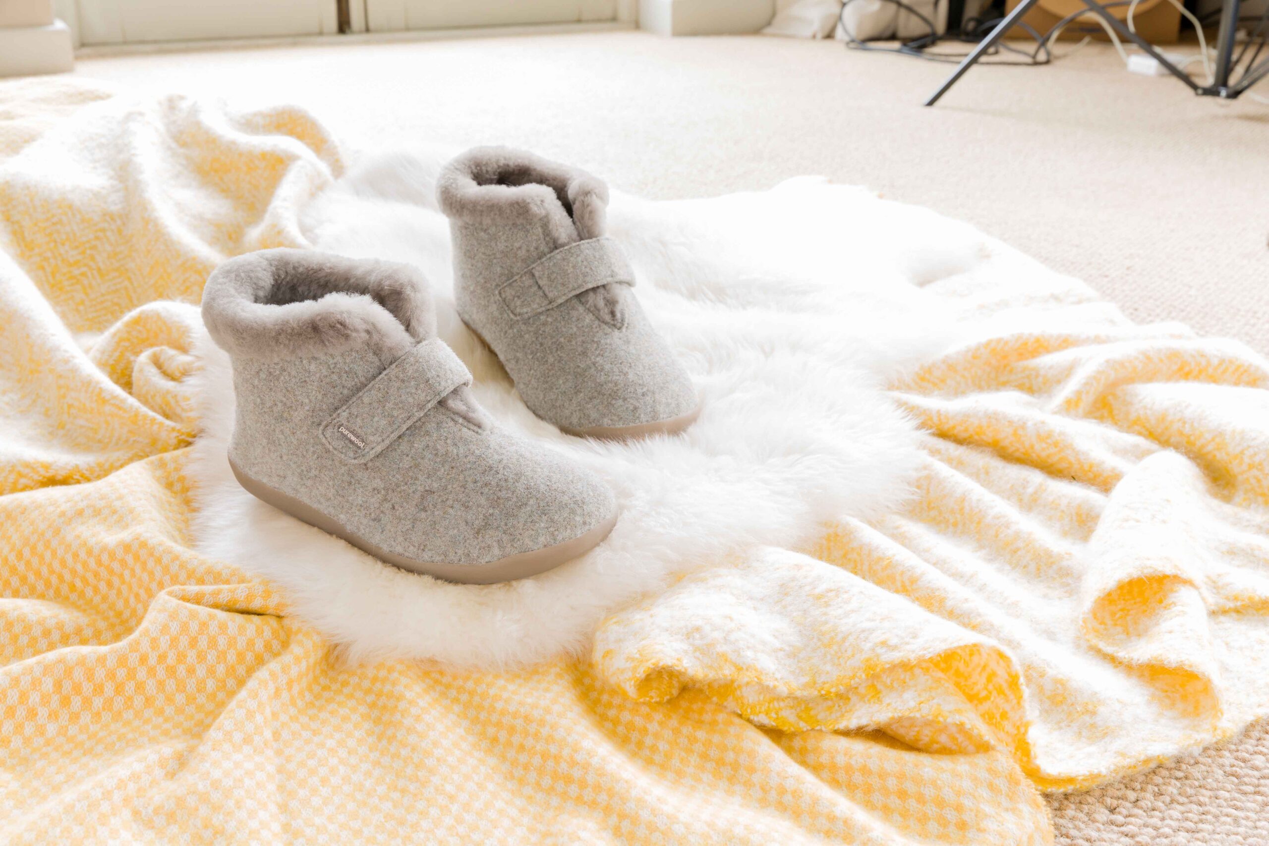 Cosyfeet Anna gray slippers with fluffy lining sit on a soft white rug, surrounded by a yellow and white checkered blanket on a beige carpet, evoking warmth and comfort.
