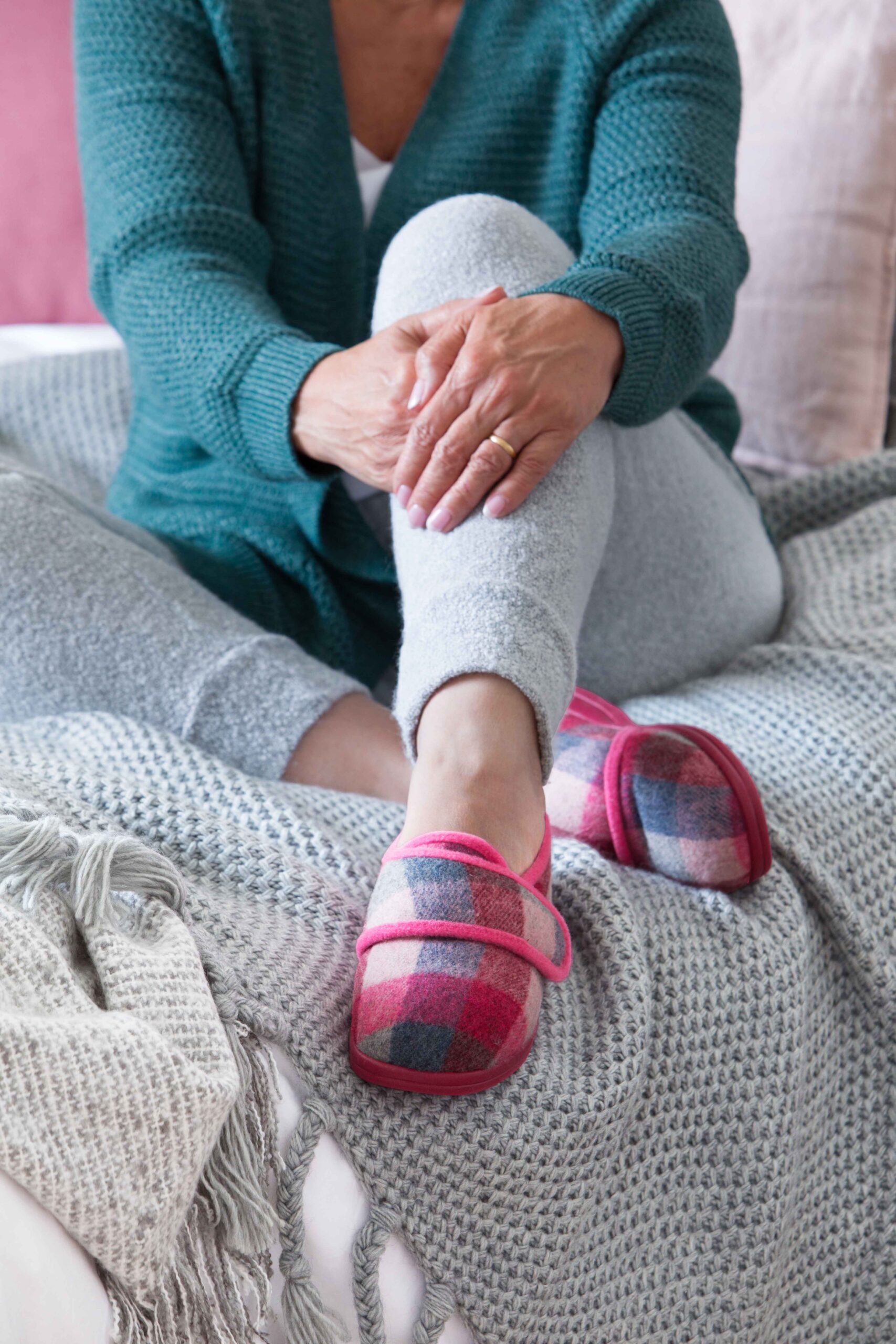 A person sits relaxed on a bed with a grey knit blanket, wearing a teal sweater, grey pants, and pink plaid Cosyfeet Holly slippers, conveying a cozy, calm mood.