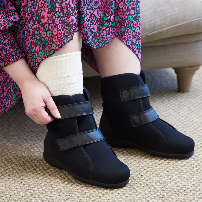 A woman in a floral dress, with a bandage on her right foot, putting on the Cosyfeet Patty in black, showcasing the flexibility of the shoe.