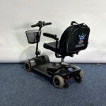 Approved Used Pride Go Go Elite Traveller in grey, with 12 ah batteries and mesh basket sitting on a navy blue carpet, behind is a white brick wall.