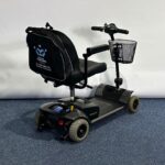 Approved Used Pride Go Go Elite Traveller in grey, with 12 ah batteries and mesh basket sitting on a navy blue carpet, behind is a white brick wall.