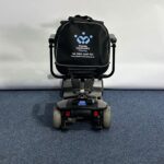 Approved Used Pride Go Go Elite Traveller in grey, with 12 ah batteries and mesh basket sitting on a navy blue carpet, behind is a white brick wall.