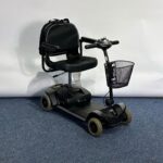 Approved Used Pride Go Go Elite Traveller in grey, with 12 ah batteries and mesh basket sitting on a navy blue carpet, behind is a white brick wall.