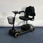 Approved Used Pride Go Go Elite Traveller in grey, with 12 ah batteries and mesh basket sitting on a navy blue carpet, behind is a white brick wall.