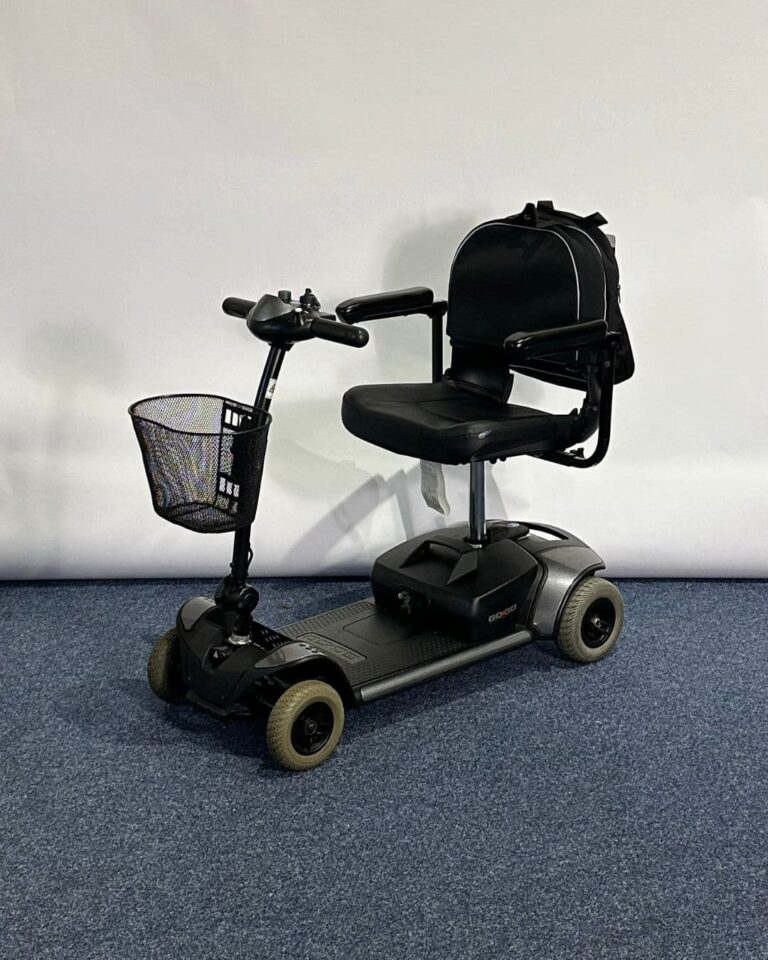 Approved Used Pride Go Go Elite Traveller in grey, with 12 ah batteries and mesh basket sitting on a navy blue carpet, behind is a white brick wall.
