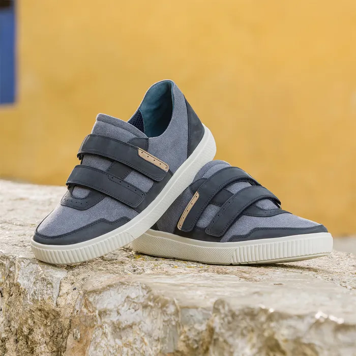A pair of stylish Cosyfeet Andrew Navy trainers with Velcro straps sits on a textured stone surface against a mustard-yellow background, evoking a modern, casual vibe.