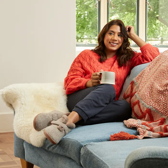 Woman in a cosy red sweater and cosyfeet Anna slippers relaxes on a blue couch, holding a mug. A patterned blanket adds warmth to the serene scene.