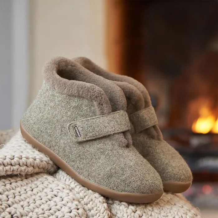 Cosyfeet Anna Pebble grey wool slippers with a Velcro strap rest on a knit blanket. A warm, softly glowing fireplace is blurred in the background, creating a warm ambiance.