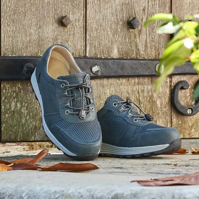 A pair of Cosyfeet Misty in Sage sitting on the group up against a vintage wooden door, one stood up leaning on the door, autumn leaves sitting on the ground around the trainers.