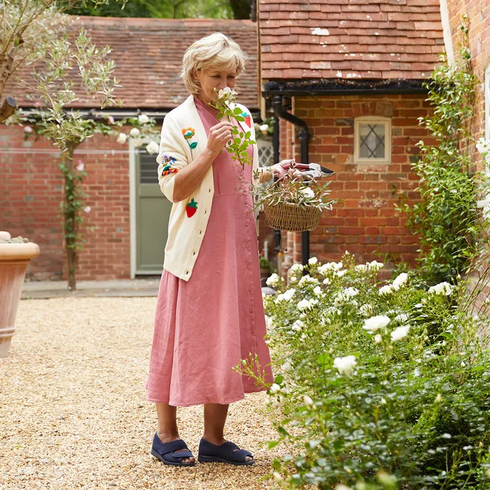 A woman in a pink dress, floral cardigan and Cosyfeet Mollie in Dark Denim, smells white flowers in a garden. She holds a basket with gardening shears, standing on a gravel path by a brick house.