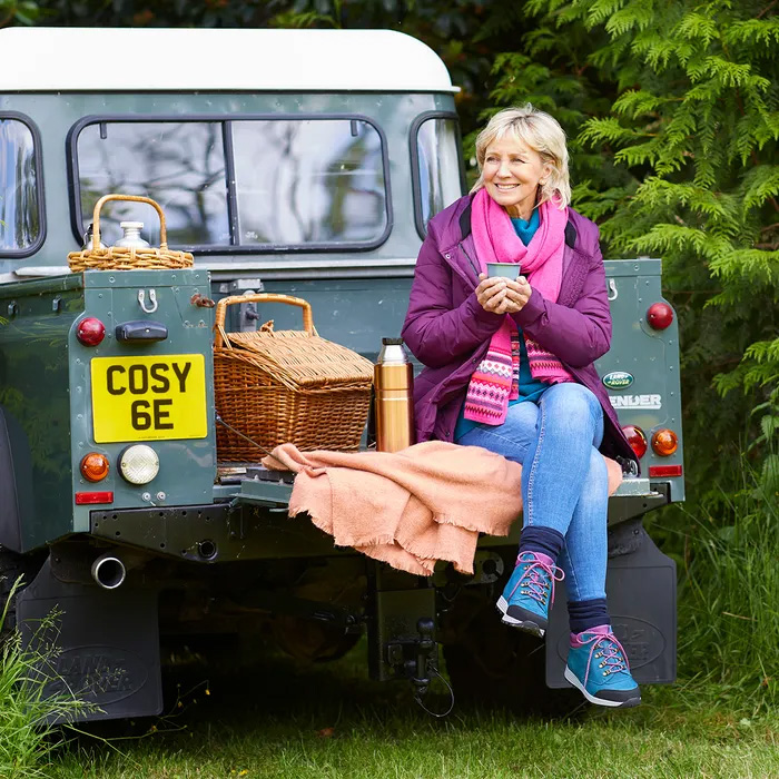 A woman in a purple coat, blue jeans and wearing Costyfeet Moose High-Performance in Teal Multi sits on the back of a vintage green car, the number plate spelling out Cosy 6E holding a mug and smiling. Nearby are a picnic basket, a flask, and a peach blanket. The scene is surrounded by lush greenery, conveying a cosy outdoor atmosphere.