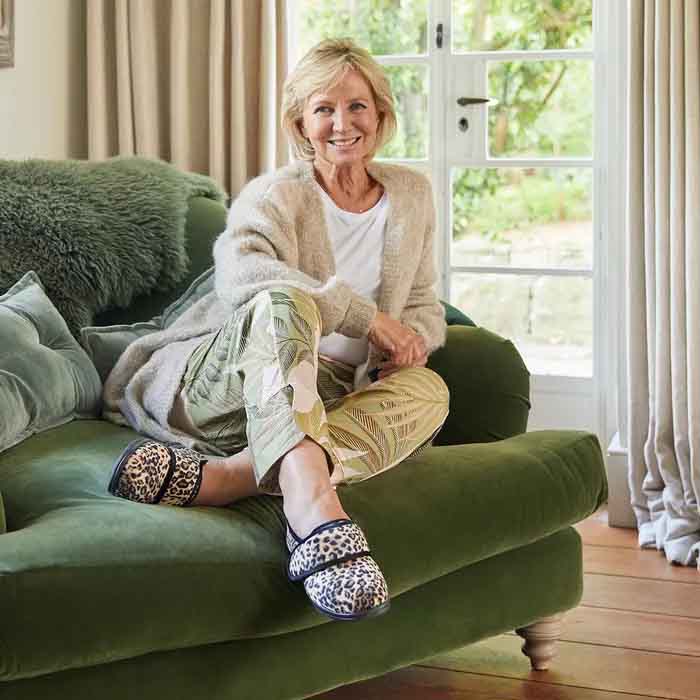 Smiling woman sits on a green sofa, wearing a cozy cardigan, patterned pants, and Cosyfeet Holly leopard plush print slippers. Bright room with large window and curtains.