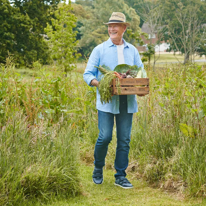 Man in a straw hat and casual outfit, wearing Cosyfeet Andrew Navy Trainers, holding a wooden crate of fresh vegetables. He's walking through a lush garden, smiling and looking content.