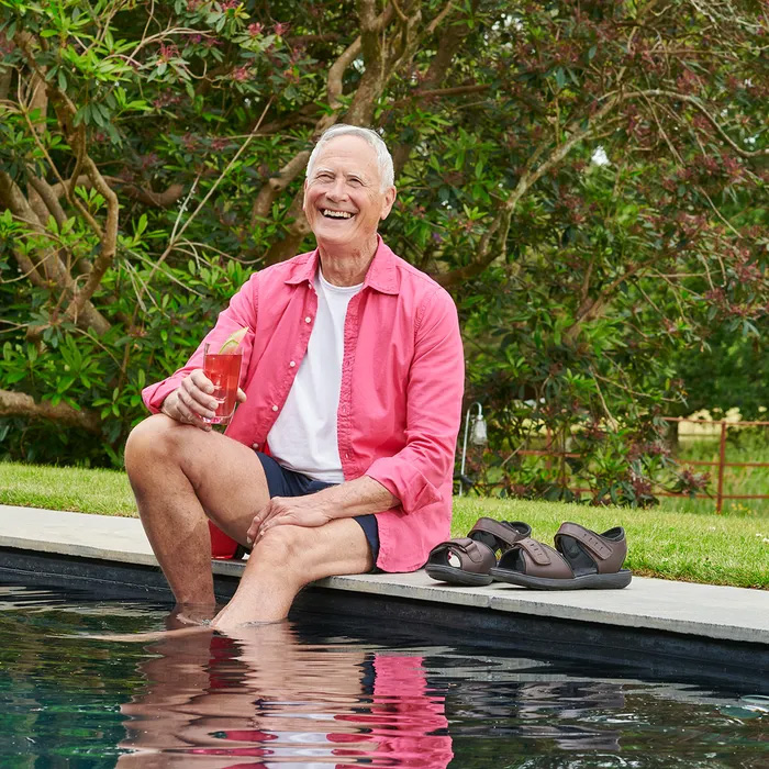 A smiling elderly man in a pink shirt sits by a pool, dipping his feet in the water, holding a red drink. Cosyfeet Bruno Brown Waxy Sandals are beside him. Lush greenery surrounds.