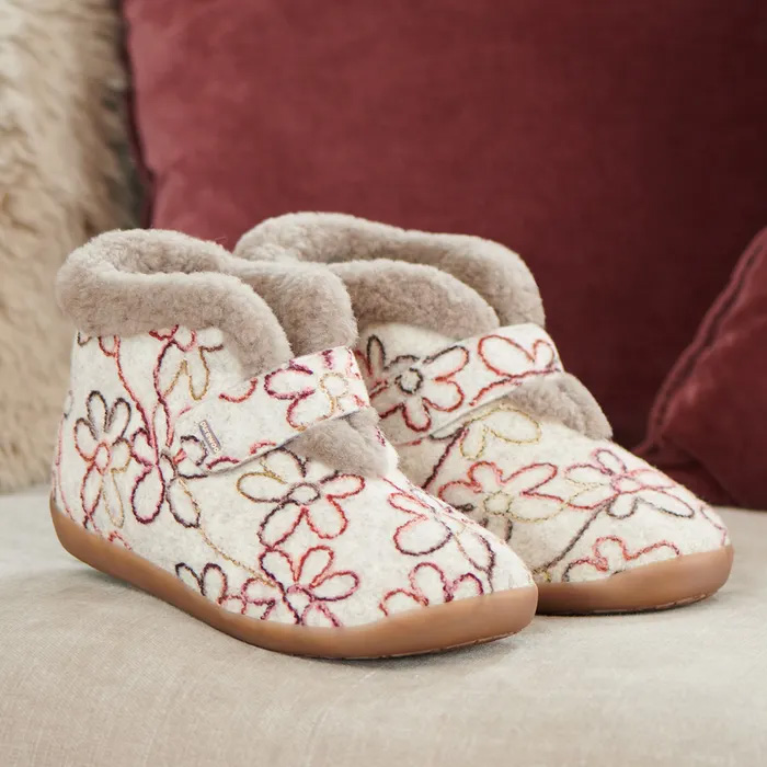 Cosyfeet Anna Wheat Floral Slippers beige slippers with soft fur lining and colorful floral embroidery, resting on a light fabric surface. Background features plush burgundy cushions.
