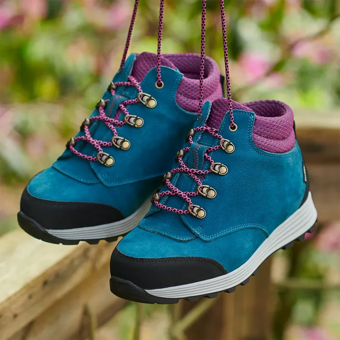 A pair of Cosyfeet Moose High-Performance in Teal Multi handing by the laces with a blurred floral background.