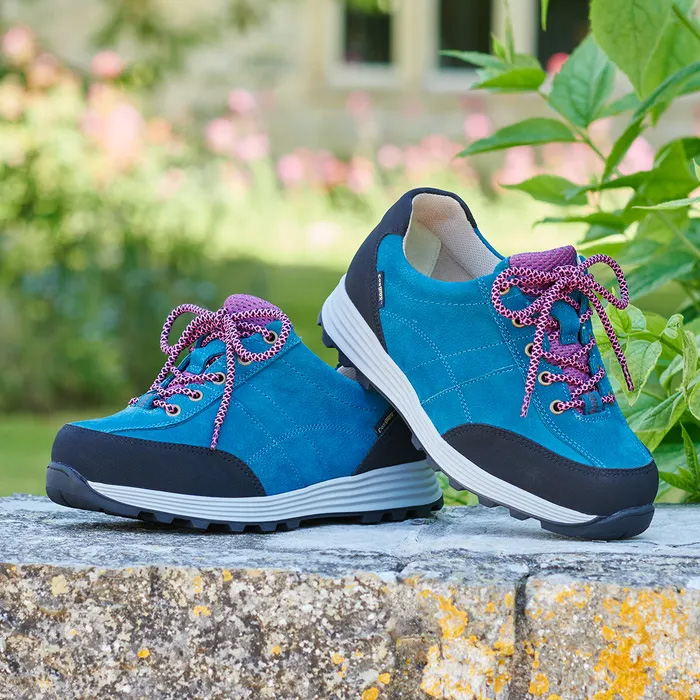 A pair of Cosyfeet Minnie High-Performance in Teal Multi sitting on a stone wall, with a blurred background of greenery and some distant windows.