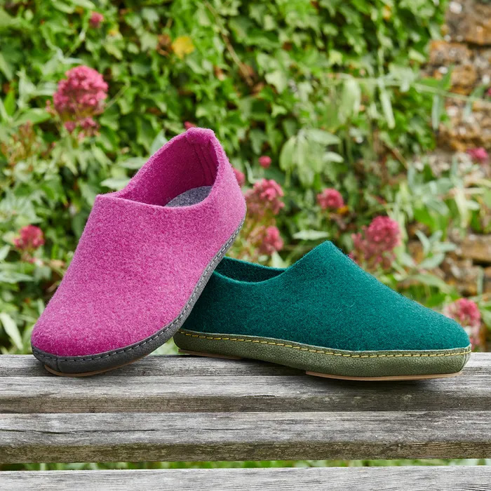 Two shoes both of Cosyfeet Mooch, one in Damson Wool and the other in Jade sitting on a wood bench in front of a floral bush.