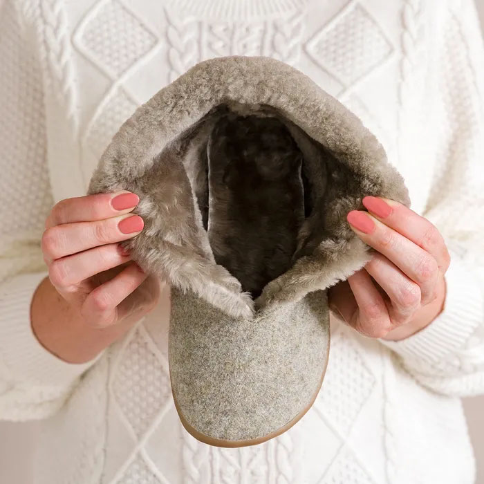 Hands holding a Cosyfeet Anna Pebble Slipper, gray faux-fur-lined slipper. The soft texture contrasts against a white, knitted sweater, conveying warmth and comfort.