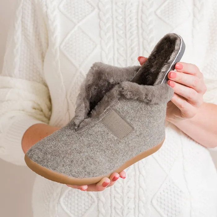 Hands holding a Cosyfeet Anna Pebble Slipper, gray faux-fur-lined slipper. The soft texture contrasts against a white, knitted sweater, conveying warmth and comfort. Showing the sole.