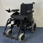 Quickie Rumba Powerchair sitting on a blue carpet and white brick wall.