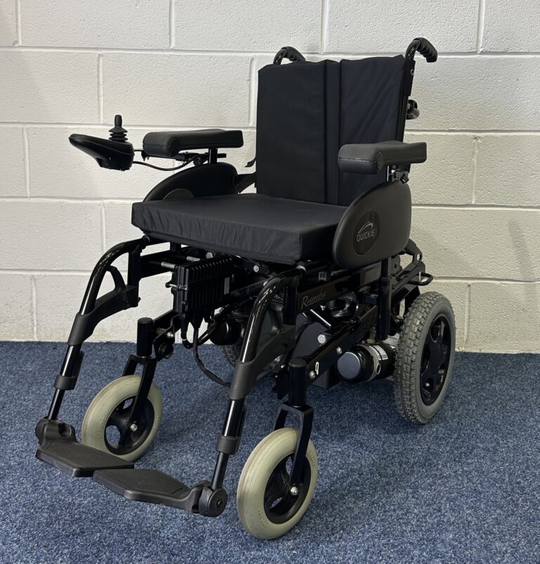 Quickie Rumba Powerchair sitting on a blue carpet and white brick wall.