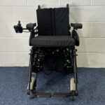Quickie Rumba Powerchair sitting on a blue carpet and white brick wall.