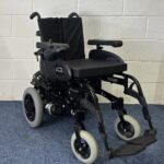 Quickie Rumba Powerchair sitting on a blue carpet and white brick wall.