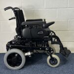 Quickie Rumba Powerchair sitting on a blue carpet and white brick wall.