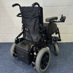 Quickie Rumba Powerchair sitting on a blue carpet and white brick wall.
