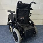 Quickie Rumba Powerchair sitting on a blue carpet and white brick wall.