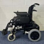 Quickie Rumba Powerchair sitting on a blue carpet and white brick wall.