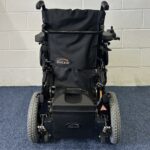 Quickie Rumba Powerchair sitting on a blue carpet and white brick wall.