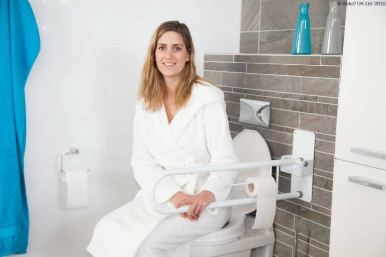A woman sitting on a toilet in a dressing robe holding onto the Atlantis Fold Away Grab Rails