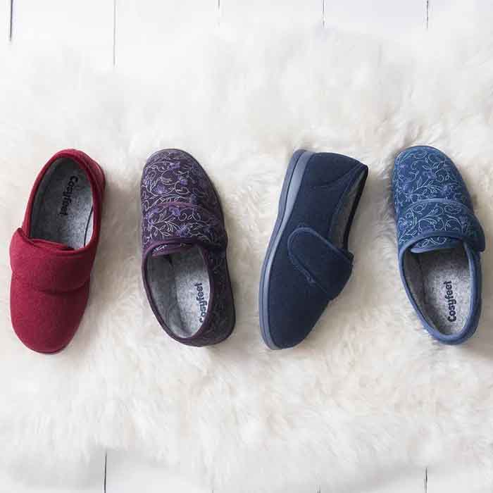 Four cosyfeet Holly slippers on a fluffy white rug. From left to right: red, purple patterned, solid navy, and light blue patterned. Warm and inviting.