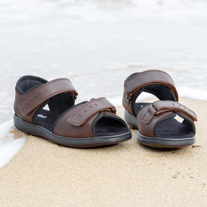 Cosyfeet Bruno Brown Waxy Sandals sitting on the sand of the beach with the water from the sea washing around them.