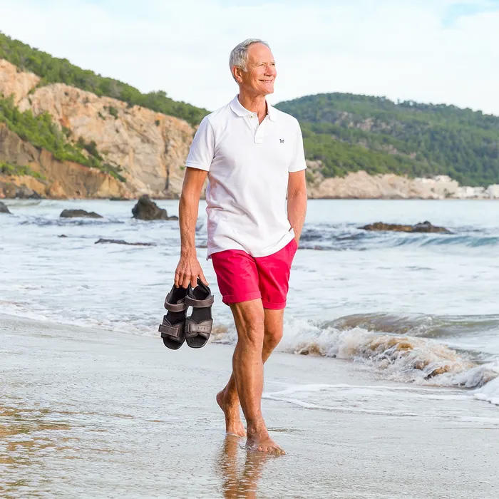 An older man smiles while walking barefoot on a sandy beach, holding Cosyfeet Bruno Brown Waxy Sandals. He wears a white polo and red shorts. Cliffs and waves are in the background.