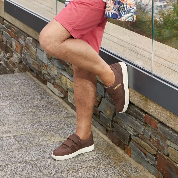 A person wearing red shorts and Cosyfeet Andrew Chocolate trainers leaning against a stone wall. The mood is relaxed, highlighting casual summer style.