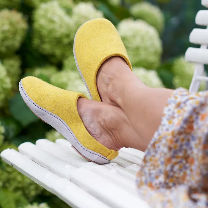 A woman's feet up on a bench wearing Cosyfeet Mosey in Yellow, with a floral dress just showing to the right hand side of the image, behind the bench is a greenery from a garden bush.