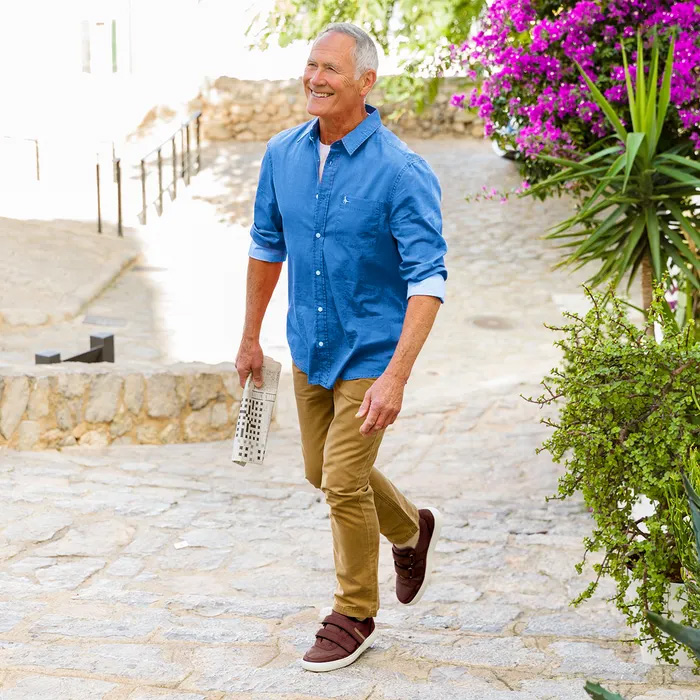 Man in a blue shirt and khaki pants with Cosyfeet Andrew Chocolate Trainers, smiles while walking up a stone path, holding a newspaper. Bright pink flowers and greenery are in the background.