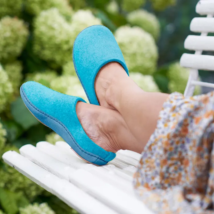 A woman's feet up on a bench wearing Cosyfeet Mosey in Turquoise, with a floral dress just showing to the right hand side of the image, behind the bench is a greenery from a garden bush.