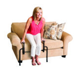 A woman wearing a pink top and white trousers pushing herself up from a cream sofa using the Stander EZ Stand -N- Go.