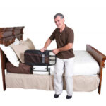 A man sitting on the side of a bed in a brown top and white trousers leaning on the Stander EZ Adjustable Bed Rail