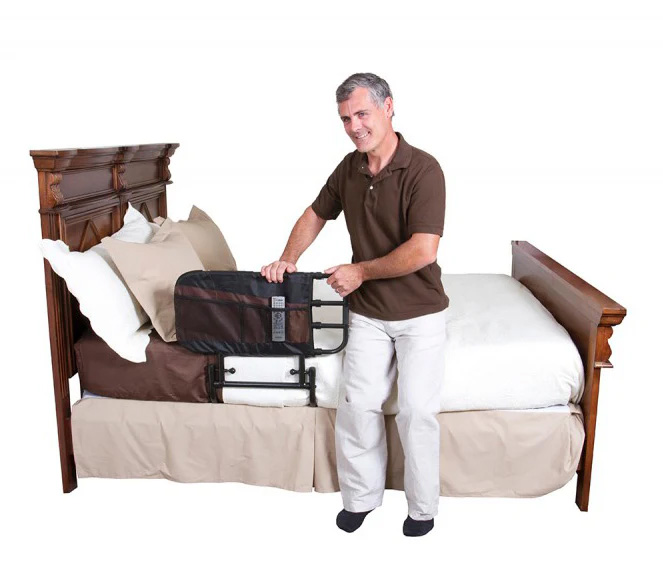A man sitting on the side of a bed in a brown top and white trousers leaning on the Stander EZ Adjustable Bed Rail