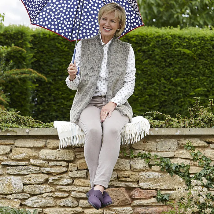 A woman sits on a stone wall, smiling under a polka dot umbrella. She wears a fur vest and Cosyfeet Spicy in Loganberry and holds the umbrella above her. Green hedges are in the background.