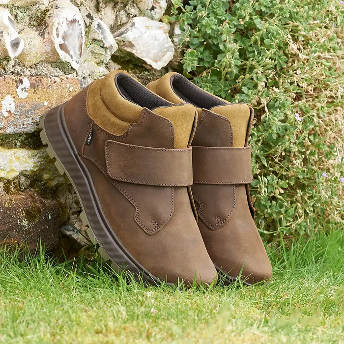 Cosyfeet Scott High-Performance Highland Boots with Velcro straps are leaning against a stone wall, surrounded by green grass and ivy. They convey a rustic feel.