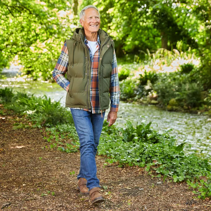 A senior man strolls along a leafy forest path by a serene stream, wearing a green vest and plaid shirt and Cosyfeet Scott High-Performance Highland boots, exuding contentment and relaxation.